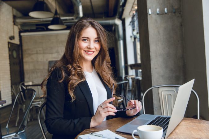 Widely smiling businesswoman working on laptop sitting in a cafe. Woman with long hair wears black jacket and white blouse and holds black eyeglasses in hands.